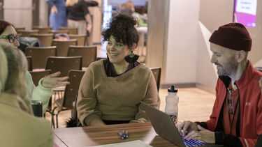 Four people sat having a discussion around a table while someone takes notes on a laptop.