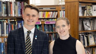 Two people stand next to one another and smile at the camera, and in front of them in a cabinet are artefacts from a museum exhibition.