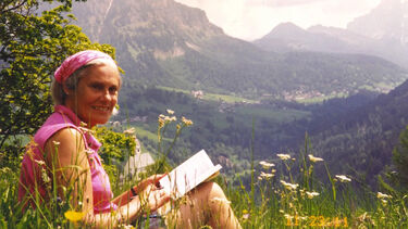 A more recent shot of Maureen sitting with a book in a grassy field overlooking a mountainscape, smiling and wearing a pink headscarf