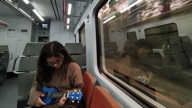 Girl on an empty train with a blue ukulele. She is looking down but we can see she is wearing glasses