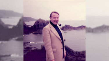 Arthur standing against a backdrop of the sea and rugged cliffs, wearing a suit jacket and smoking a pipe
