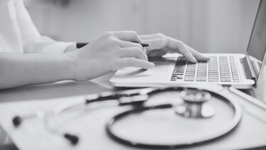 A person works at a laptop with a stethoscope visible in the foreground.