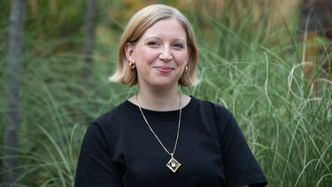 Image of woman with a blonde bob smiling, wearing a black top and gold necklace with greenery in the background