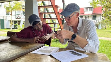 Two men sitting at a table looking at a document that one is taking a photo of