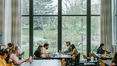 Four people sat at a table using laptops to study. Trees and park greenery show in the background