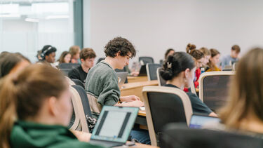 Person sat at a desk typing at a laptop surrounded by others also attending the lecture