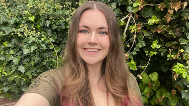 Amy has long brown hair with pink ends. She wears a green top and stands in front of a hedge.