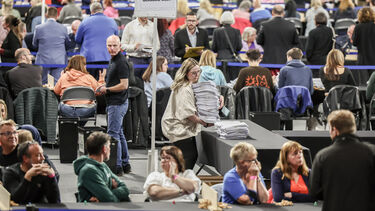 Lots of people in a room, sat around tables, whilst a person brings a stack on election ballots 
