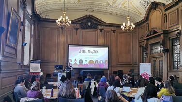 A woman stood at a podium in a wood panelelled hall talking to an audience seated at tables.