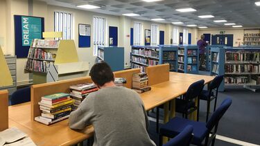 A library with a student sat at a desk and several shelves of books