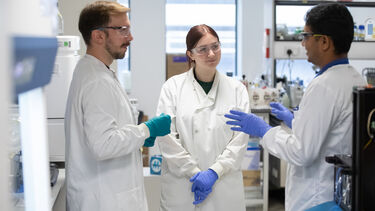 Three researchers, wearing white lab coats, having a discussion in their lab