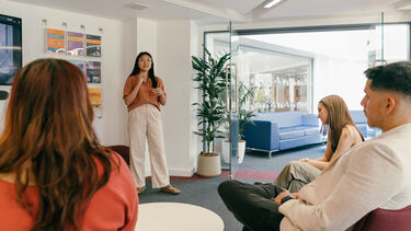 A woman in glasses standing up presents to three seated onlookers