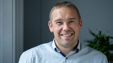 A photo of Dave smiling, looking relaxed and friendly. He has short, cropped brown hair and is wearing a blue shirt. He's in a grey room, and in th background there is a plant.