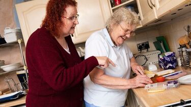 A woman supports an older woman to prepare food in the kitchen 