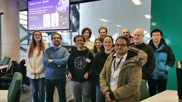 A small group photo in a computer room against the background of a large interna glass and a large screen displaying details of the Insigneo/Sano Modelathon