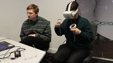 Two men sitting at a desk, one is wearing a metaquest headset and holding the controllers