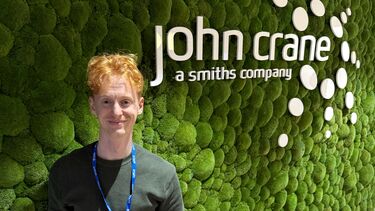 A young man stands in front of a green wall with the text 'John Crane UK'