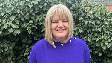 Laura has a blond bob and a blue jumper. She stands in front of some greenery.