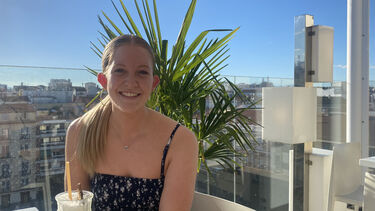 Sophie has her blonde hair in a ponytail and sits on a terrace in front of a palm tree and blue sky
