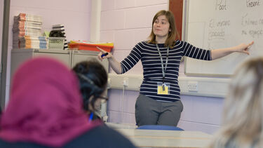 A female teacher stands at the front of a seminar room, pointing to a whiteboard. In the foreground, there are the heads and shoulders of students, who are listening to what the teacher says.