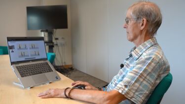  Gemini said A man in a plaid shirt and glasses sits at a wooden table, looking at a laptop screen that displays a cognitive assessment with four illustrated panels. He is using a computer mouse, and a small microphone is clipped to his shirt.