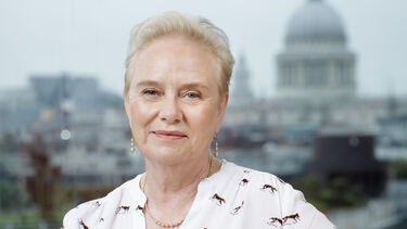 Woman with cropped blonde hair, dressed smarly in a white patterned shirt sat confidently on a yellow sofa
