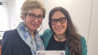Two women standing next to each other, holding a sheet of paper with the University logo as a masthead