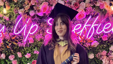 A female student in a graduation gown standing against a floral photo wall