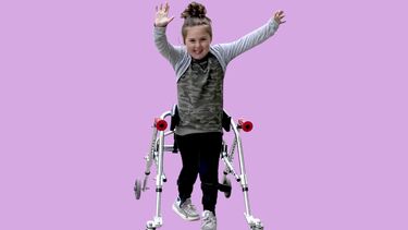 A young girl smiling and raising her arms, standing up with the assistance of a walker