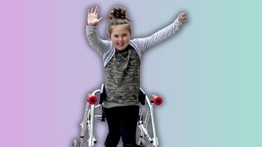 A young girl smiling and raising her arms, standing up with the assistance of a walker