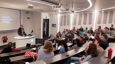 An audience in a lecture theatre looking at a screen with the film title "People's Emergency Briefing" displayed on it.