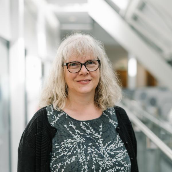 Profile picture of Carolyn Axtell wearing black framed glasses, a black cardigan and a dark grey with white pattern.