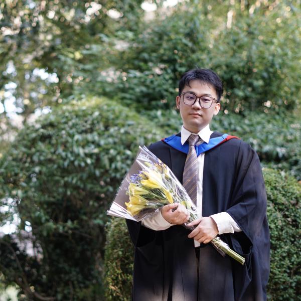 Profile picture of Xiao Cheng in a black graduation gown, holding a bunch of yellow flowers.