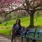 A photograph of Ifedayo Ayodeji sitting on a park bench underneath a tree with pink blossom. 