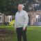 John Lavender wears a grey sweatshirt. He is stood outside an old stone building with a tree on the left.