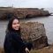 Picture of Yoana sitting outside on a grey and cloudy day, on the edge of a coastline with a view of birds nesting on isolated rocks and cliff faces. 