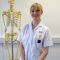 Isabel Gatenby has blonde hair tied back. She is wearing a white student midwife uniform and standing in front of an anatomical skeleton.