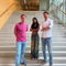 A student stood on a staircase in a University building with two academic members of staff stood either side, all of them looking at the camera.