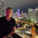 A young male stands in the foreground on a balcony and look towards the camera, resting his arm on the side of the balcony. It is night time and behind them is a brightly light cityscape with tall buildings.