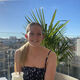 Sophie has her blonde hair in a ponytail and sits on a terrace in front of a palm tree and blue sky