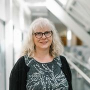 Carolyn Axtell wearing black framed glasses, a black cardigan and a dark grey with white pattern.