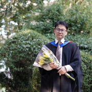Xiao Cheng in a black graduation gown, holding a bunch of yellow flowers.