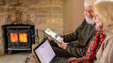 An older couple, a man and a woman sit of on a sofa using a laptop and an tablet each, a lit log burning stove is in the background