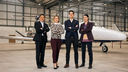 four people, one asian man, a white woman, a white man and an asian woman, stand in a hanger with a small plane in the background