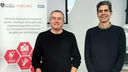 Two men in navy blue jumpers smiling in front of an Insigneo Institute bannerstand.