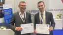 Two smiling men in grey suits holding british cardiovascular society award certificates against a background of conference banners