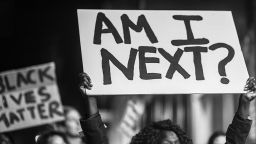 A black and white photo of a woman holding a placard reading 'Am I next?'