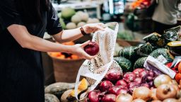 Cropped shot of young Asian woman shopping for fresh organic red onions in supermarket. She is shopping with a cotton mesh eco bag and carries a variety of fruits and vegetables. Zero waste concept