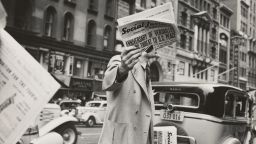A man dressed in early twentieth century clothing, stood on a street surrounded by old motor vehicles, holds a newspaper up