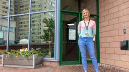 Amy Harding wears a pastel ombre jumper and skinny blue jeans. She has blonde hair. She stands in front of a glass panelled building.